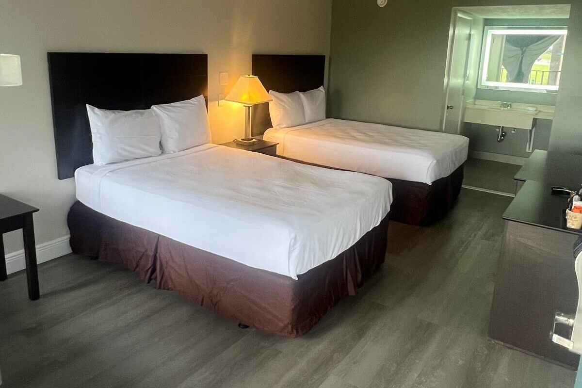 A hotel room with two beds, a nightstand with a lamp, a small table, a desk, and a mirror above the desk.
