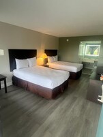 Habitación de hotel con dos camas, una mesita de noche con lámpara, una pequeña mesa, un escritorio y un espejo sobre el escritorio.
