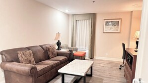 A living room with a brown leather sofa, a small table, and a chair. There is a window with curtains, a lamp, and a framed picture on the wall. - Clayton Plaza Hotel & Extended Stay (St. Louis)