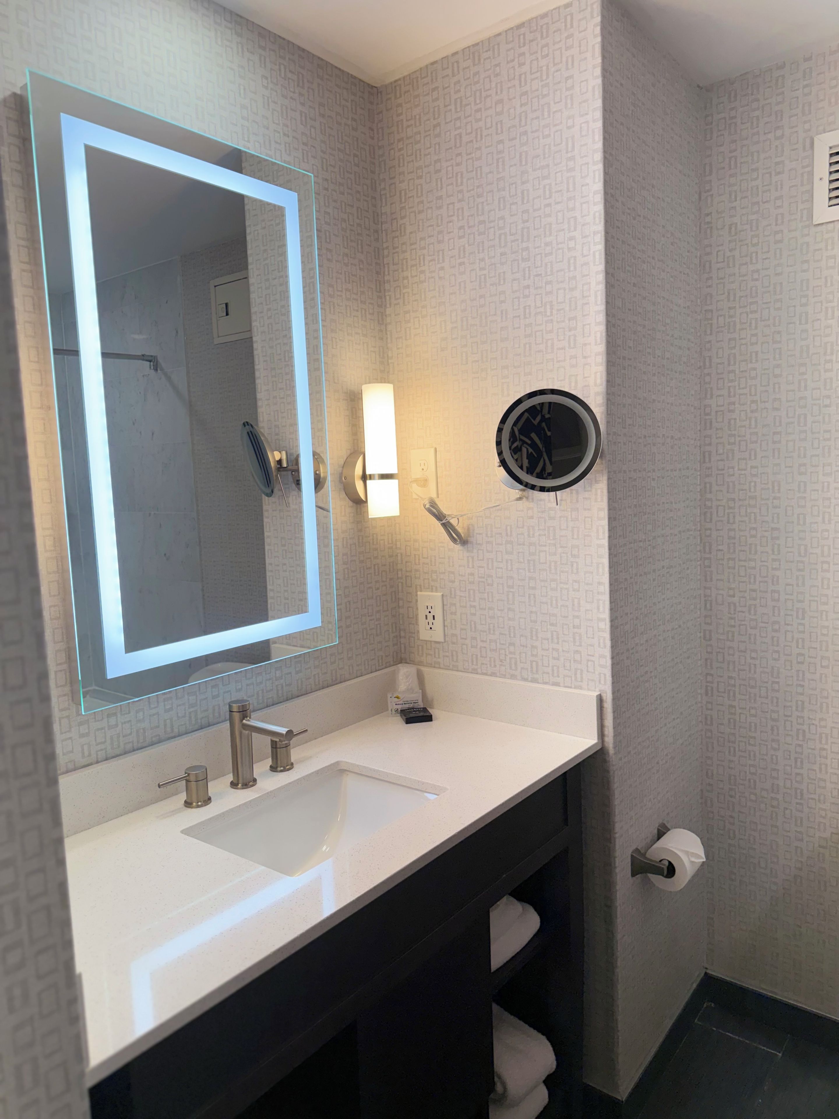 A modern bathroom with a large mirror, a white sink, a towel rack, and a hairdryer mounted on the wall.