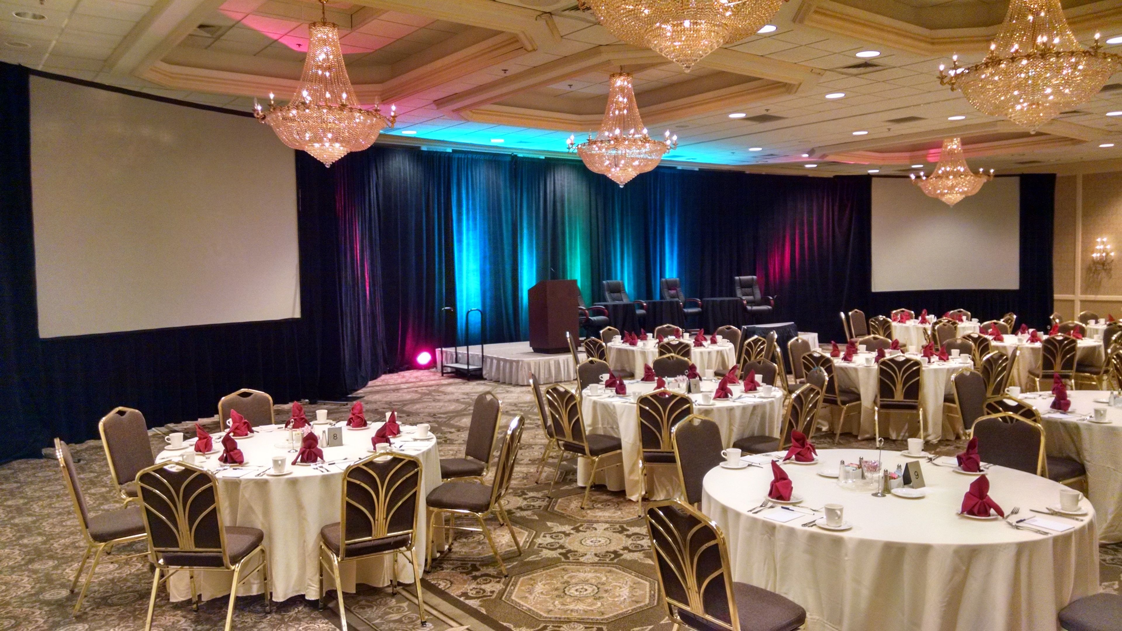 A banquet hall with round tables, gold chairs, and chandeliers.
