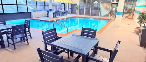 An indoor swimming pool with a tiled edge, surrounded by black chairs and tables, and a glass partition.