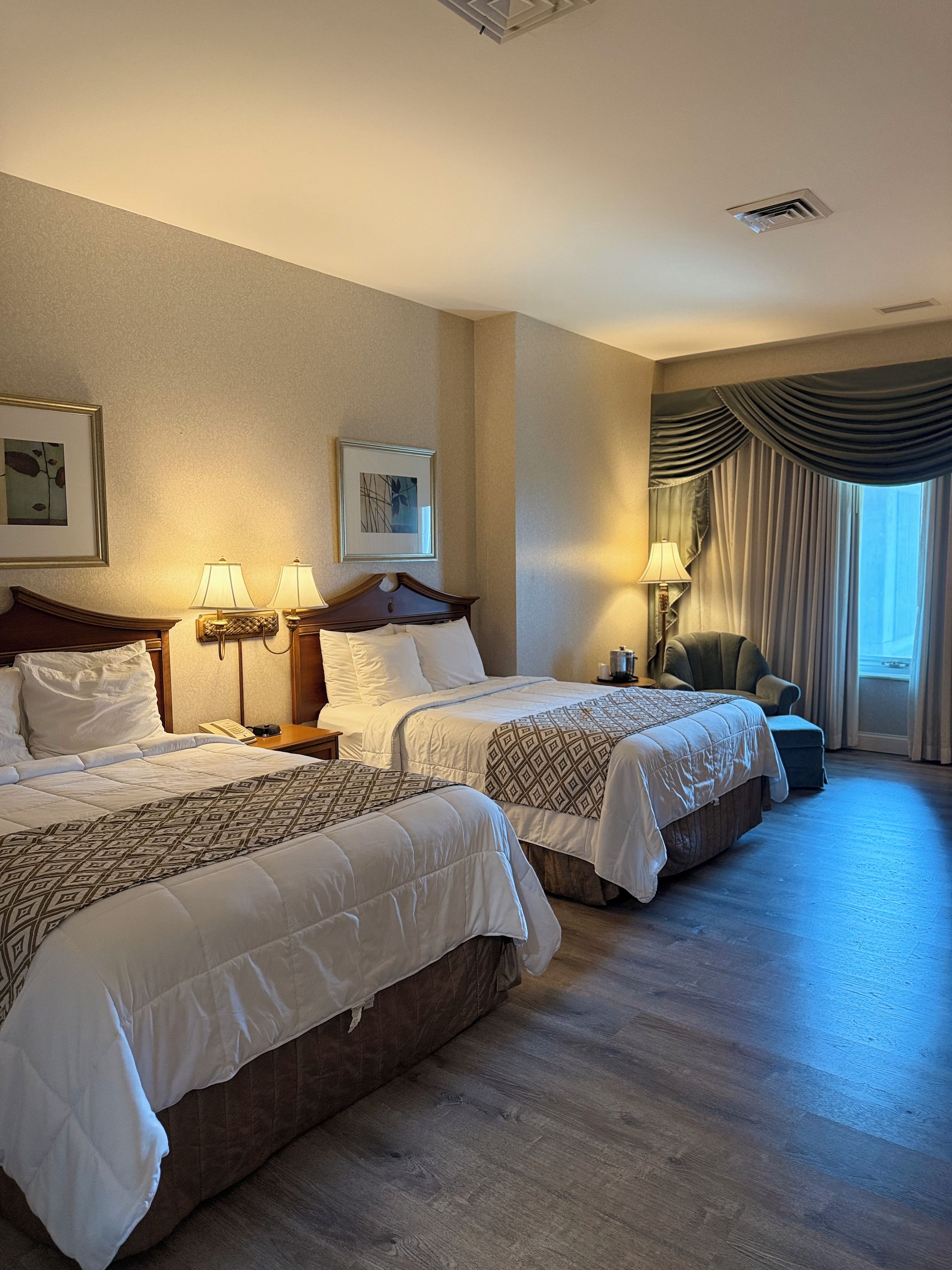 A hotel room with two beds, a sitting area with a chair, a window with curtains, and artwork on the walls.