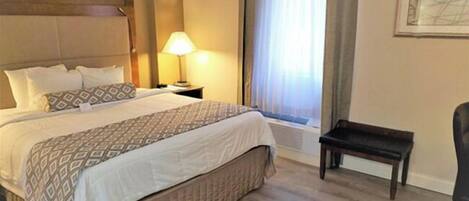 A hotel room with a bed, bedside table, lamp, window with curtains, and a chair.