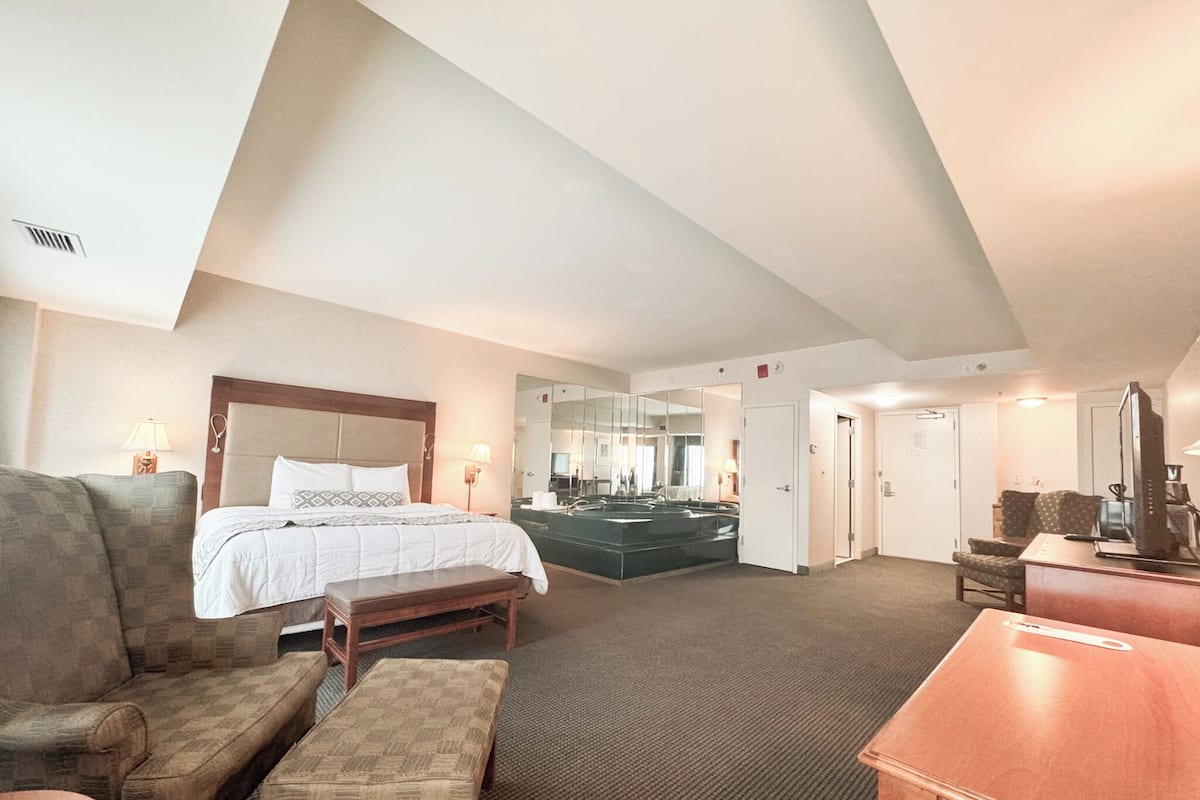 Ein Hotelzimmer mit Bett, Schreibtisch, Stuhl, Sofa, Bank, Spiegel, Badezimmer und Tür.