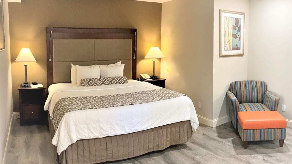 Ein Hotelzimmer mit einem großen Bett, zwei Nachttischlampen, einem gestreiften Sessel und einem orangefarbenen Hocker.
