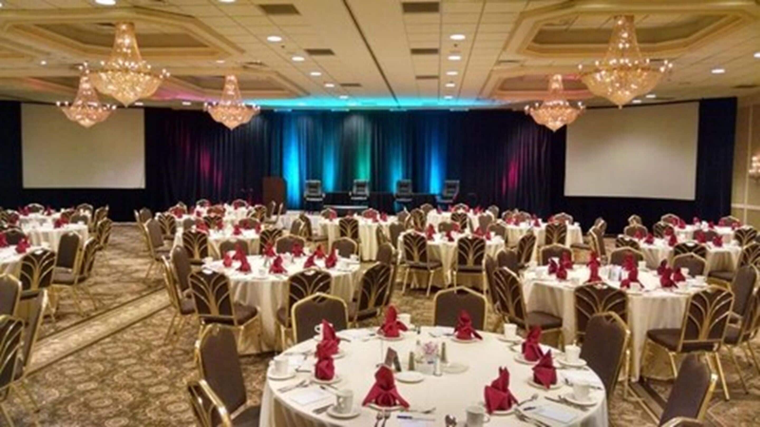 Salão de banquetes com mesas redondas preparadas para refeições, palco com fundo azul e lustres.