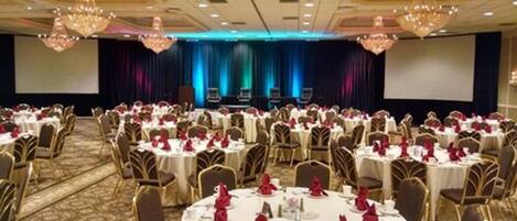 A banquet hall with round tables set for dining, a stage with a blue backdrop, and chandeliers.