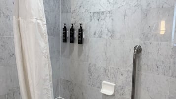 A bathroom with a glass-enclosed shower, a white bathtub, marble tiles, a handheld showerhead, and three black bottles on the wall.
