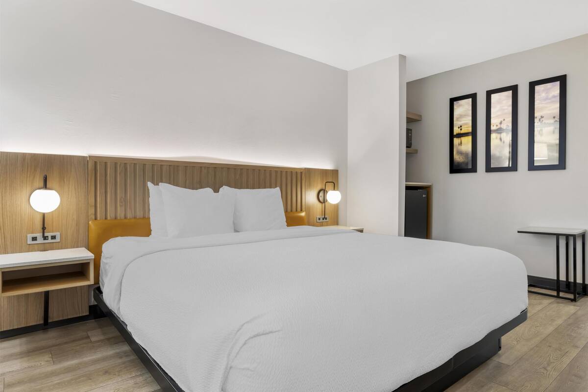 A modern hotel room with a large bed, wooden headboard, bedside tables with lamps, and a small desk with a chair.