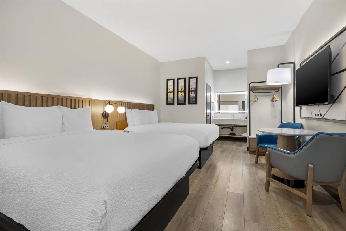 A hotel room with two beds, a desk, a chair, a TV, and a bathroom in the background.