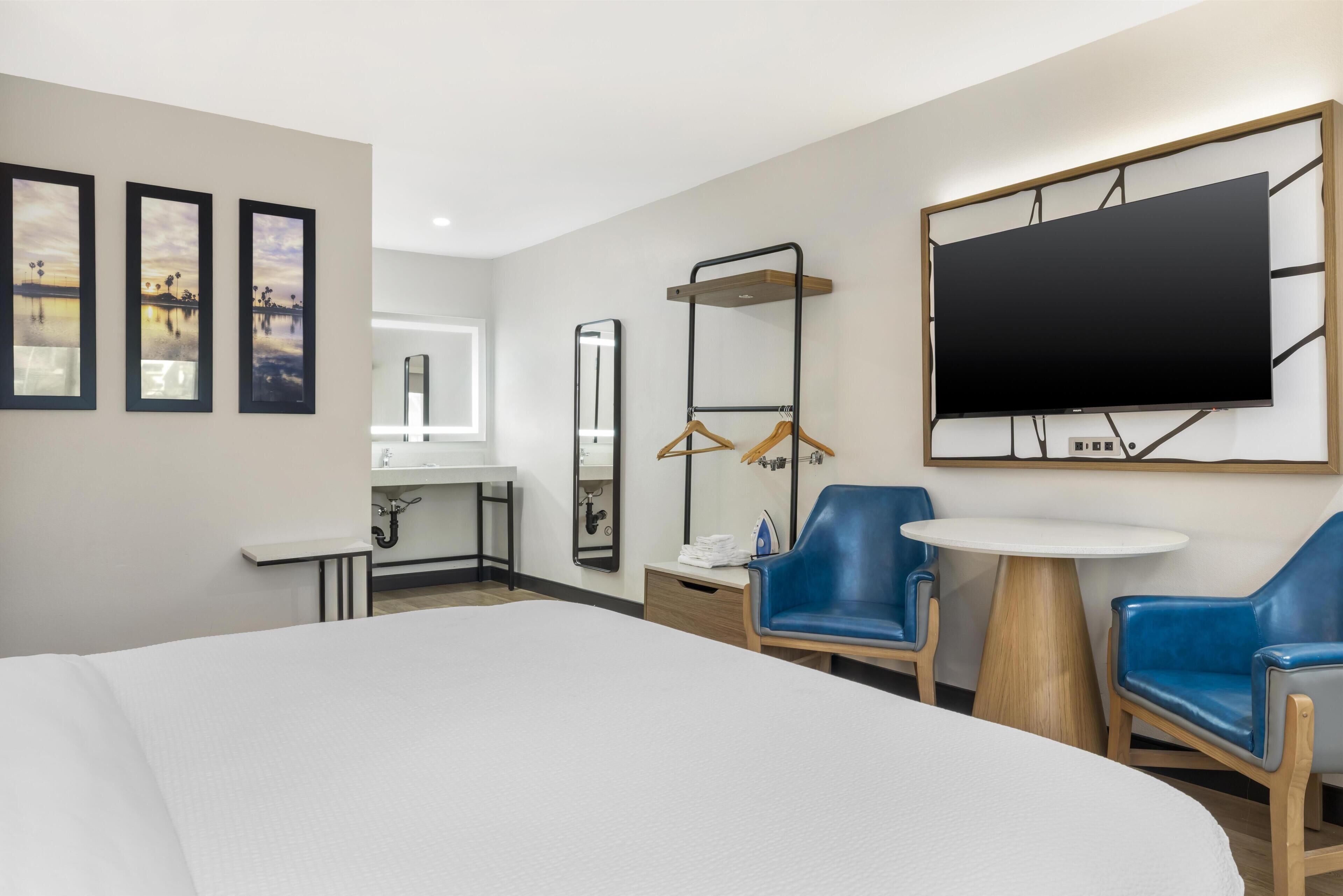 Quarto de hotel com cama, escrivaninha com espelho, televisão fixada na parede e duas cadeiras azuis.