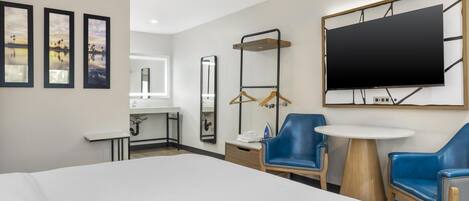 Quarto de hotel com cama, escrivaninha com espelho, televisão fixada na parede e duas cadeiras azuis.