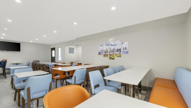 Een moderne eetruimte met tafels en stoelen, een flatscreen televisie aan de muur en een stadsgezicht kunstwerk.