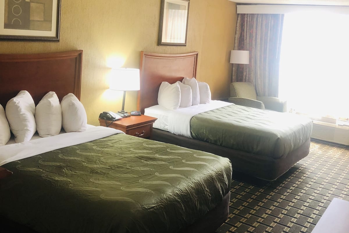 Une chambre d’hôtel avec deux lits, une tête de lit en bois, une table de chevet, une lampe et une fenêtre avec des rideaux.