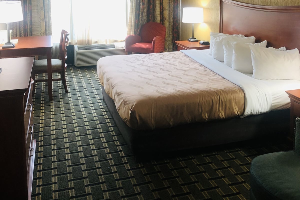 Une chambre d’hôtel avec un grand lit, un bureau, une chaise, une table de chevet, une lampe et une fenêtre avec des rideaux.