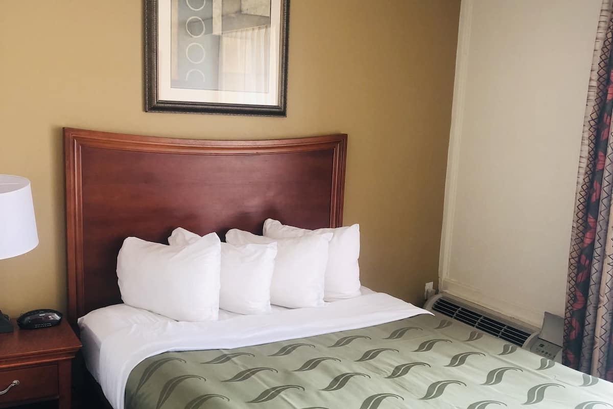 Une chambre d’hôtel avec une tête de lit en bois, un lit avec une literie blanche, une couvre-lit à motifs verts, un tableau encadré au mur, une table de chevet avec une lampe et un rideau à motifs.