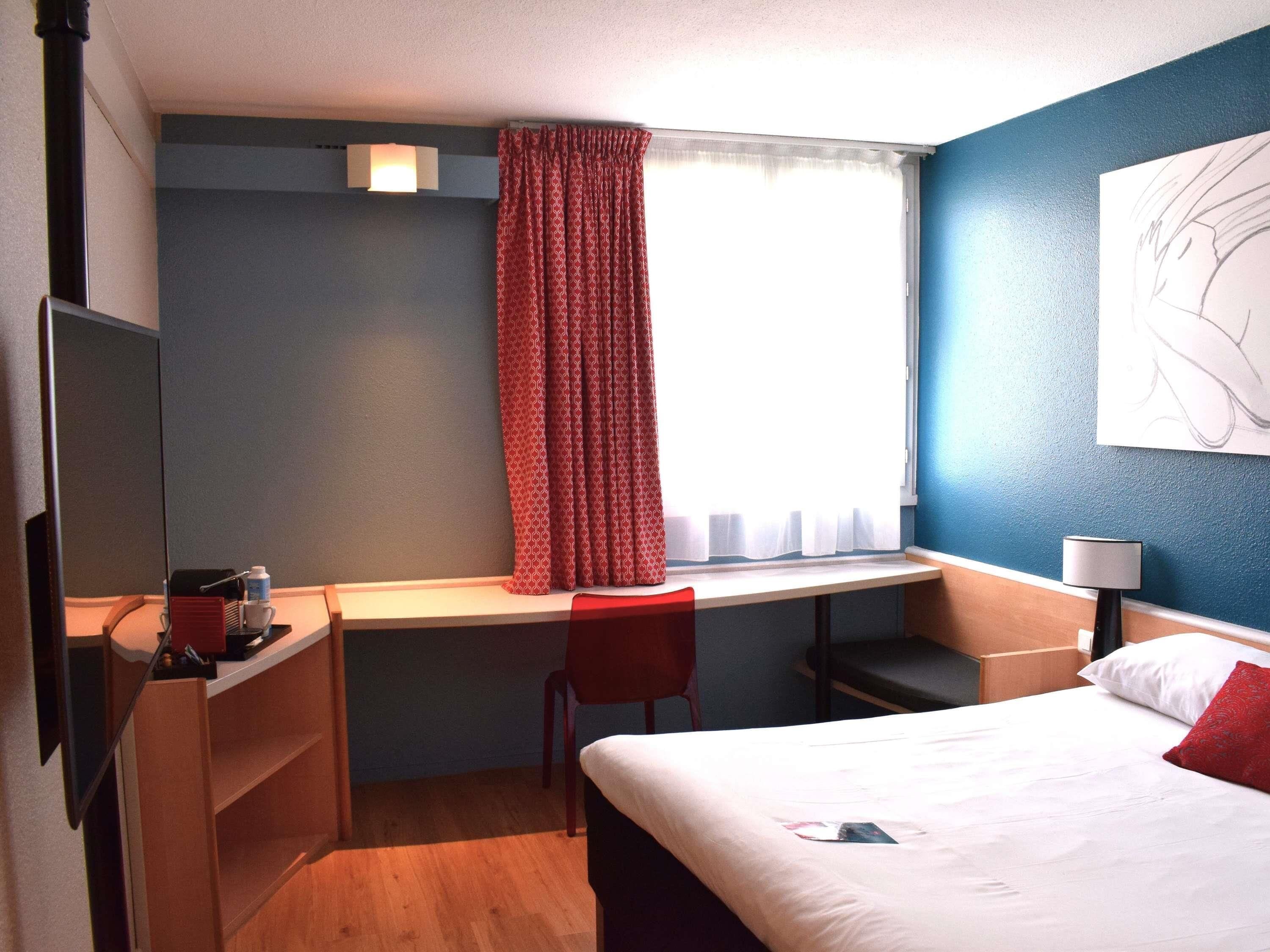 Habitación de hotel con cama, escritorio y silla, televisor, lámpara y ventana con cortinas.