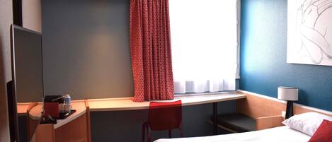 A hotel room with a bed, a desk with a chair, a TV, a lamp, and a window with curtains.