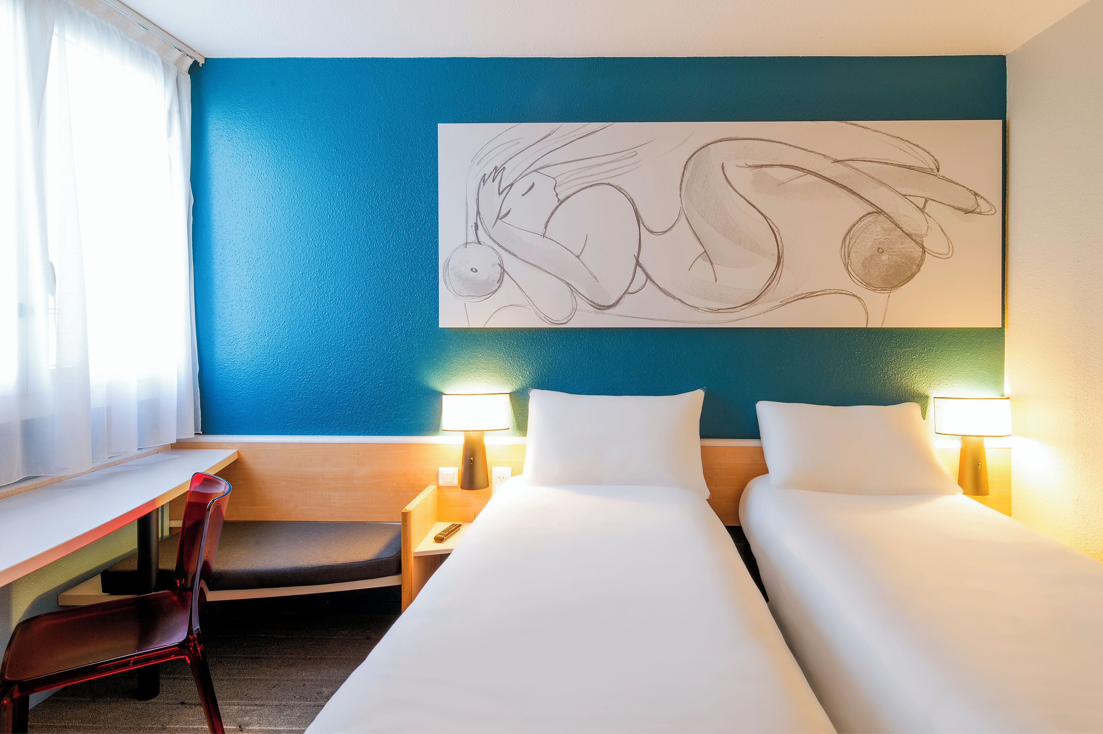 A hotel room with two beds, a desk, and a chair. There is a wall art piece above the beds and two bedside lamps.