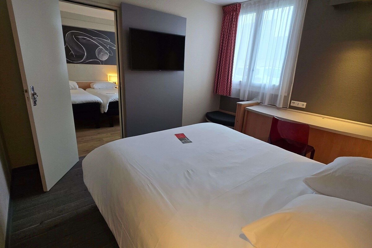 A hotel room with a bed, a desk, a TV, and a door leading to another room.