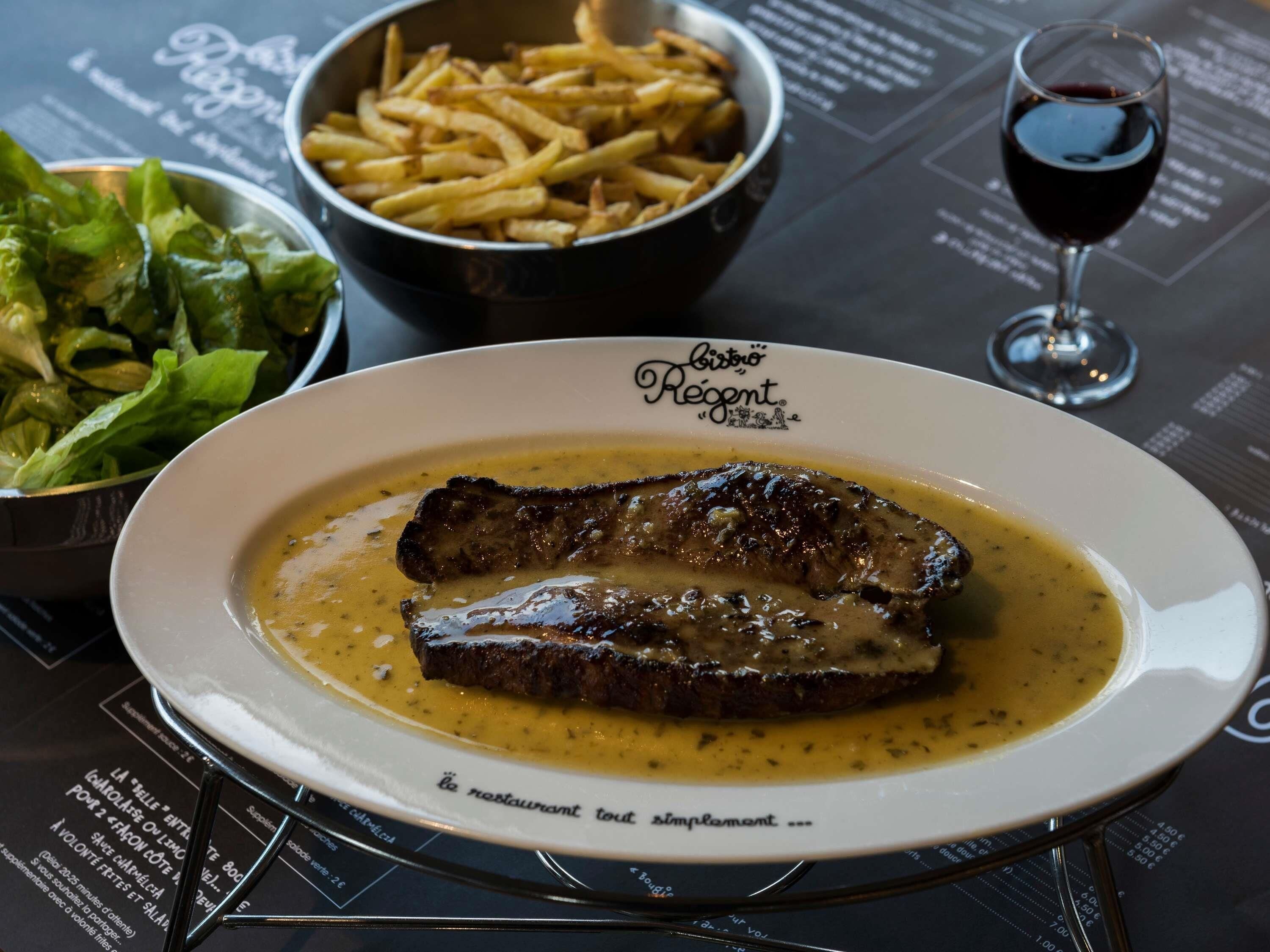 Un piatto di bistecca con salsa, una ciotola di patatine fritte, un bicchiere di vino rosso e un'insalata come contorno su un tavolo con un menu.