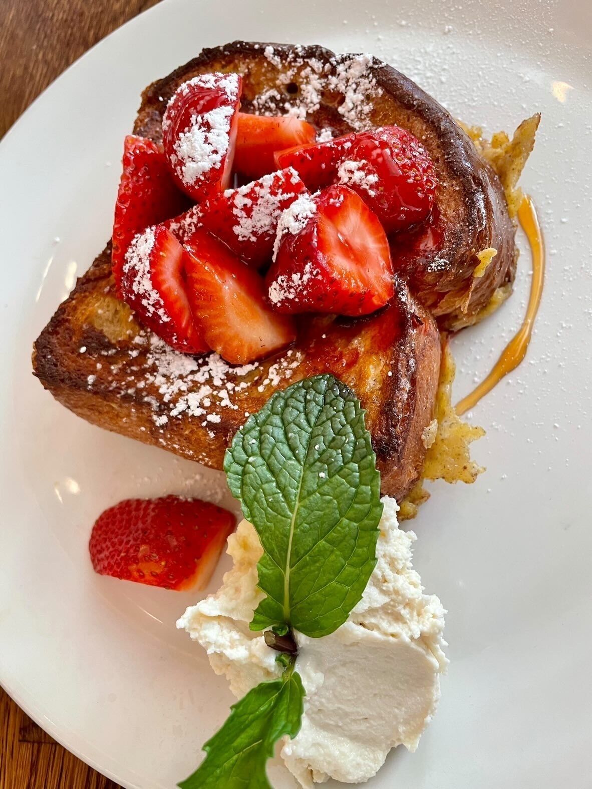 A plate with French toast topped with strawberries and powdered sugar, accompanied by a dollop of whipped cream and a mint leaf.