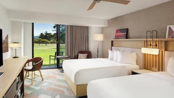 A hotel room with two beds, a desk, a chair, a ceiling fan, and a view of a grassy area.