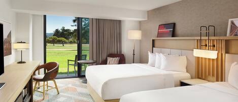 A hotel room with two beds, a desk, a chair, a ceiling fan, and a view of a grassy area.