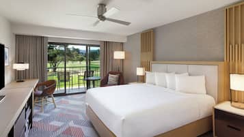 A hotel room with a large bed, a desk, a chair, a TV, a ceiling fan, and a view of a golf course.