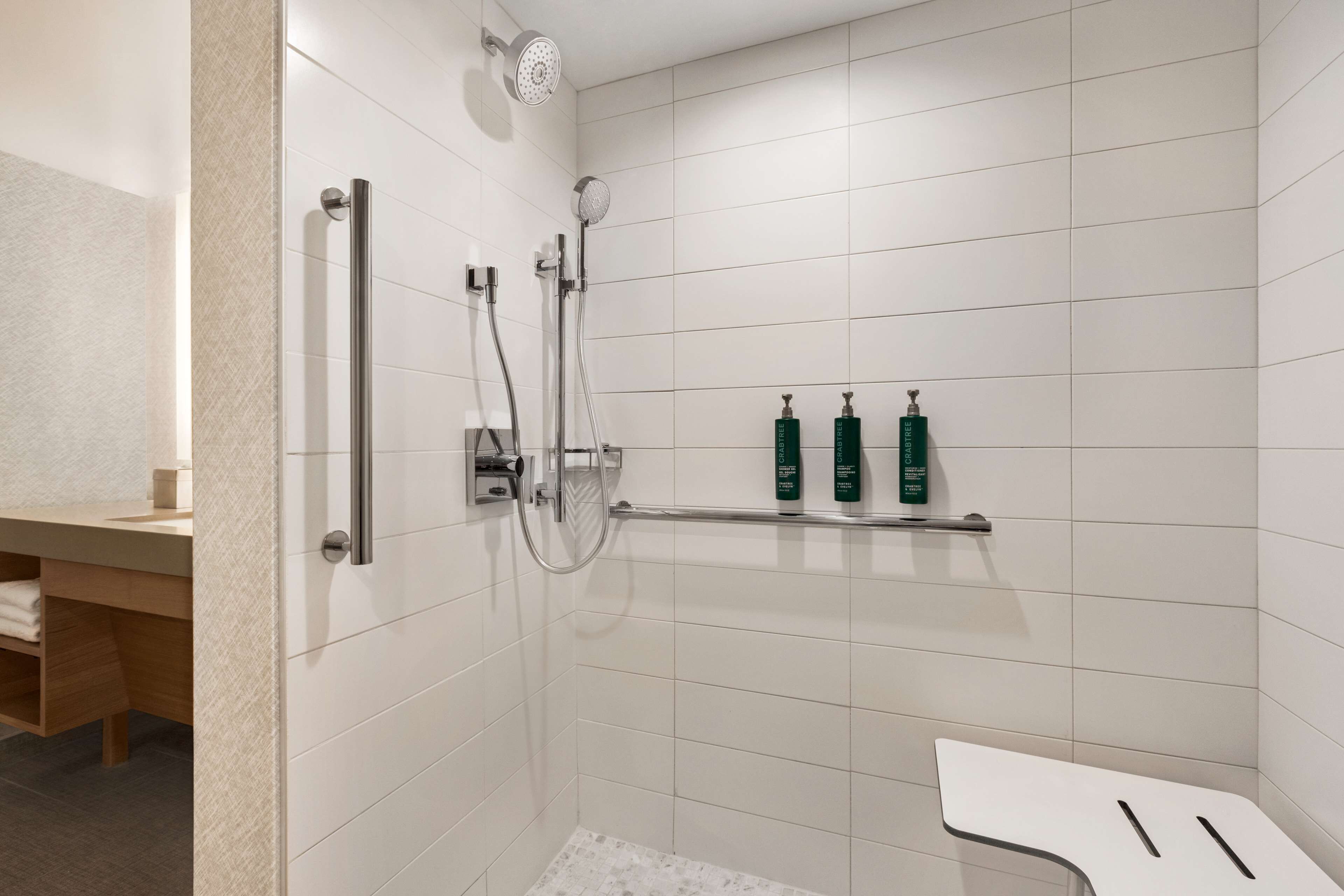Un baño moderno con baldosas blancas, una ducha con cabezal de ducha manual y un estante con botellas verdes.