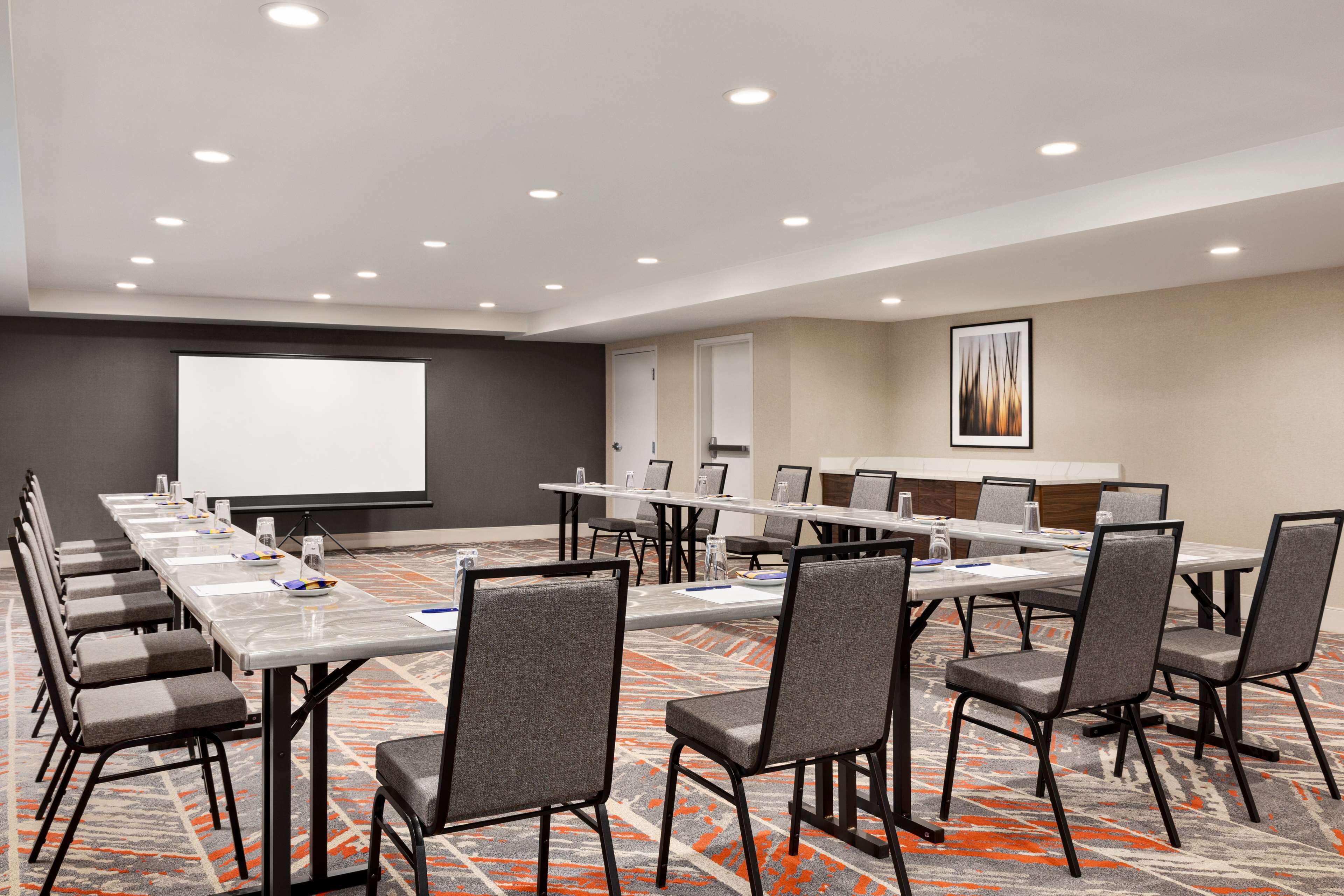 A conference room with long tables, chairs, a projector screen, and a carpeted floor.