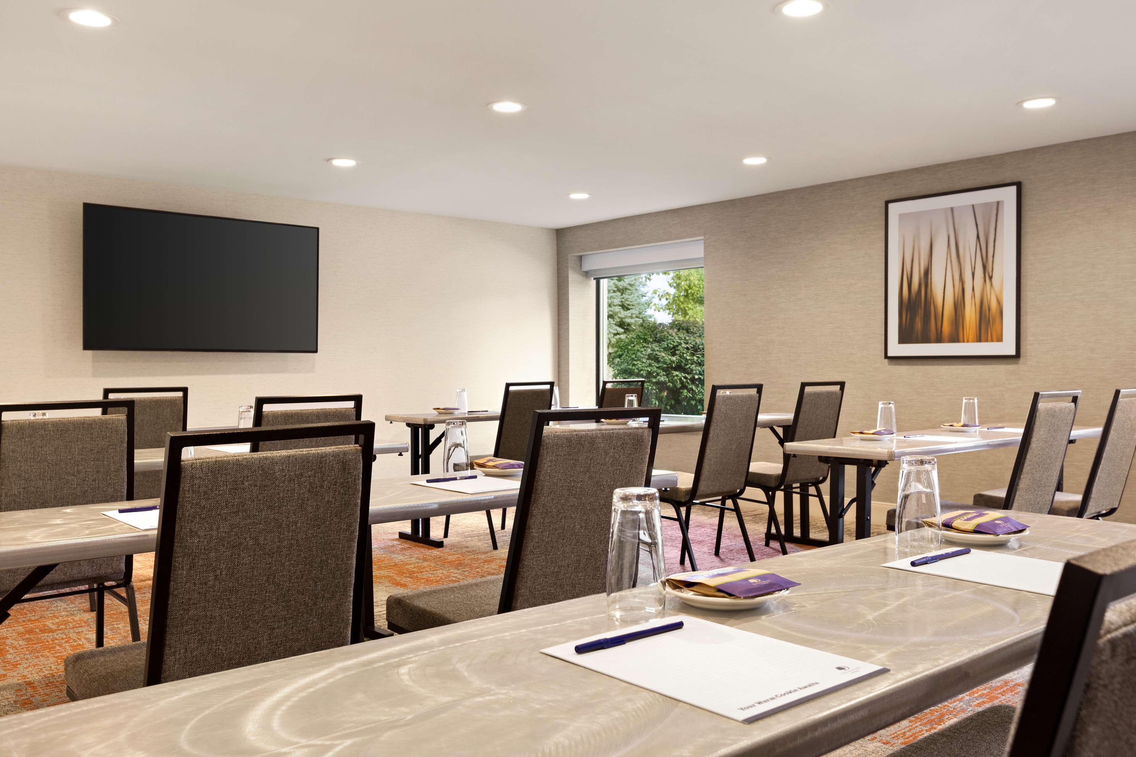 A conference room with tables, chairs, a flat-screen TV, and a window with a view of trees.