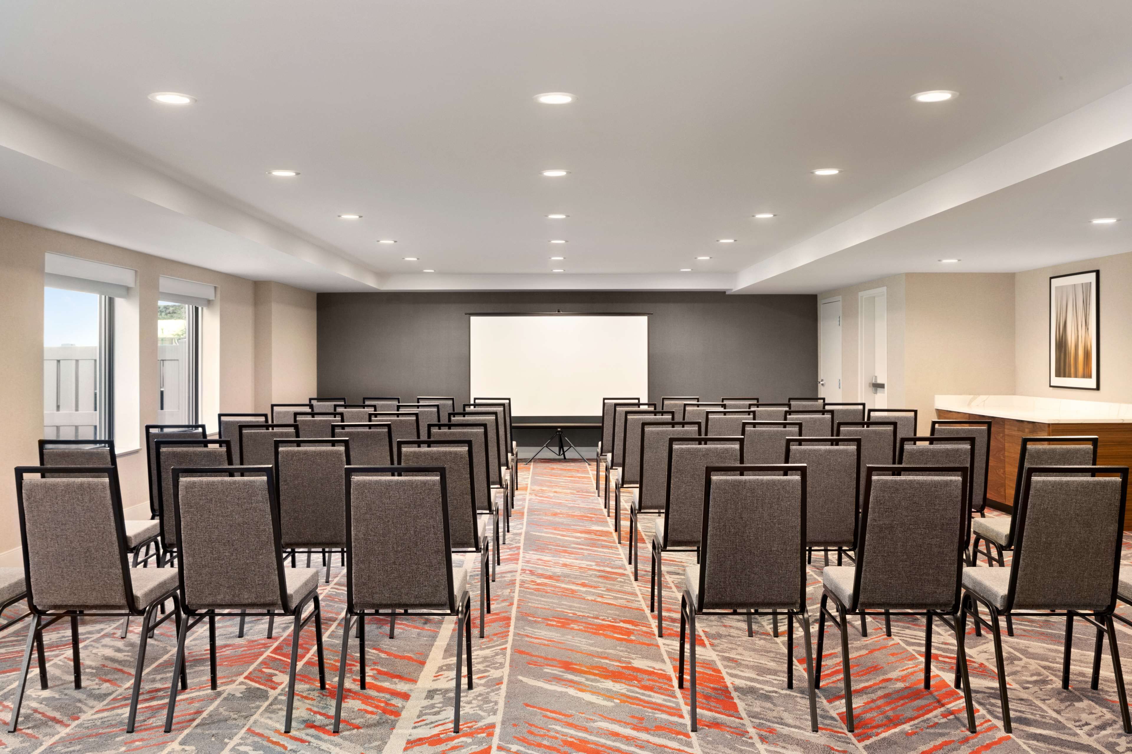 Un salón de conferencias con filas de sillas orientadas hacia una pantalla grande, piso alfombrado y un mostrador de recepción.