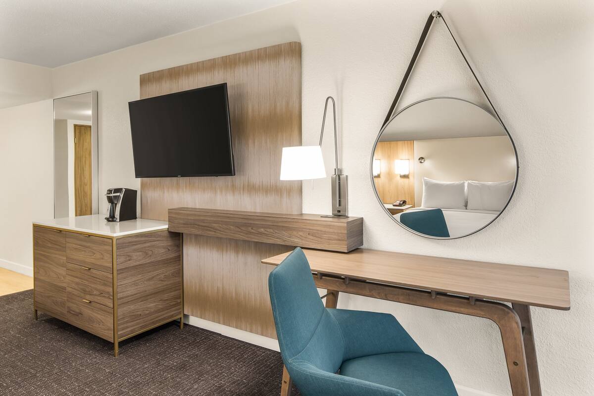 Ein Hotelzimmer mit einem Holztisch, einem blauen Stuhl, einem Flachbildfernseher, einer Lampe und einem großen Spiegel.
