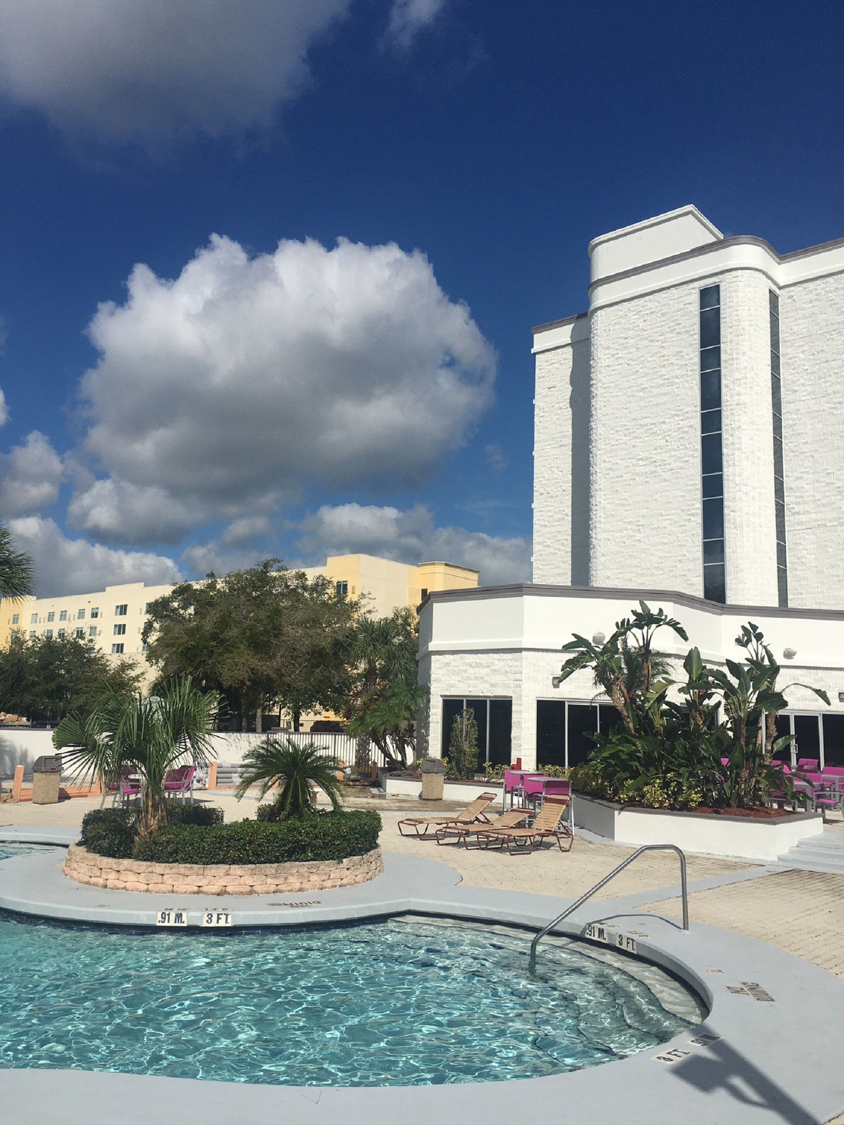 A swimming pool with a sign indicating its depth, surrounded by lounge chairs and palm trees, with a building in the background.