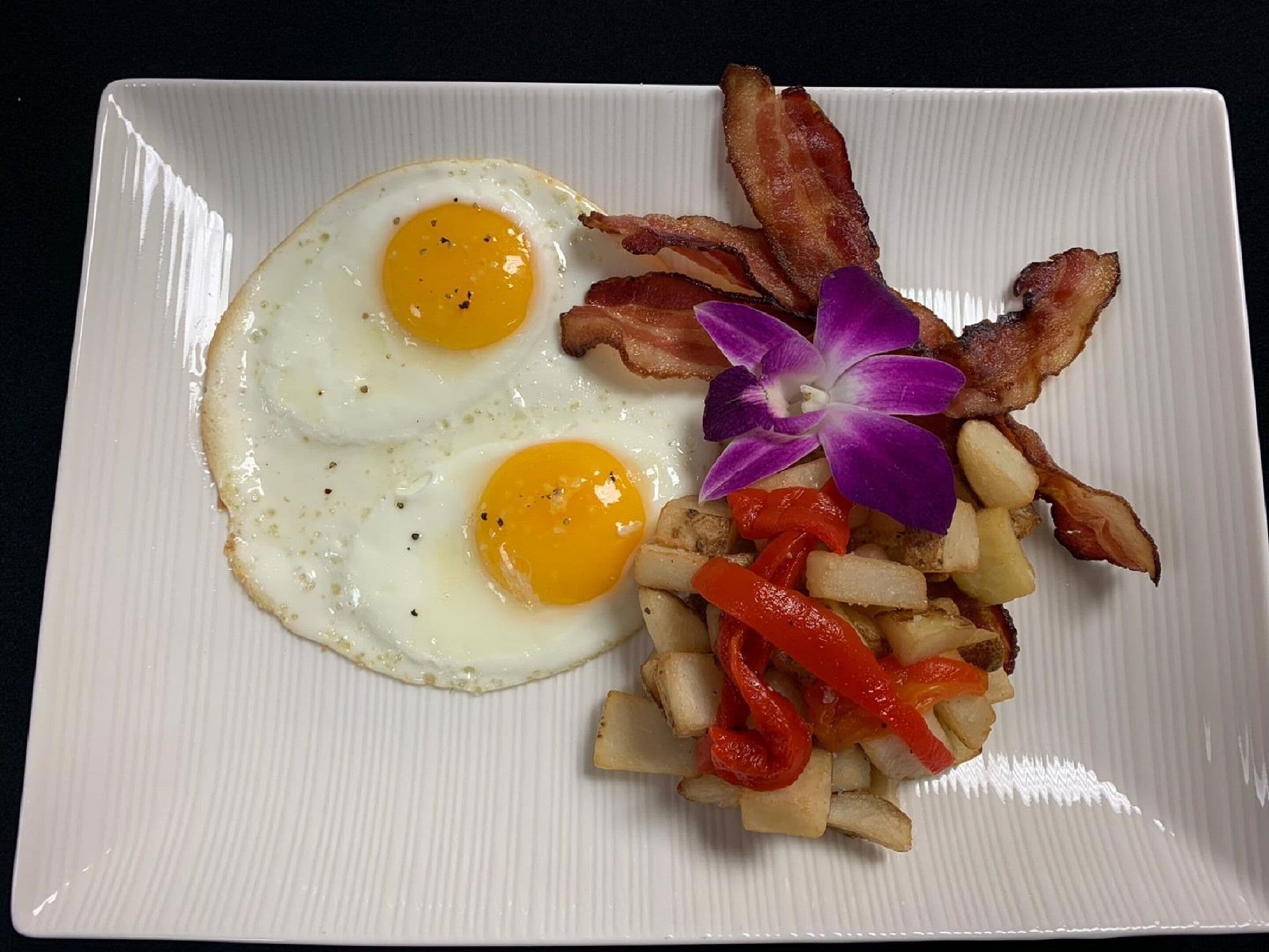 Un plato con dos huevos estrellados, tocino crujiente, patatas en cubos y pimientos rojos.