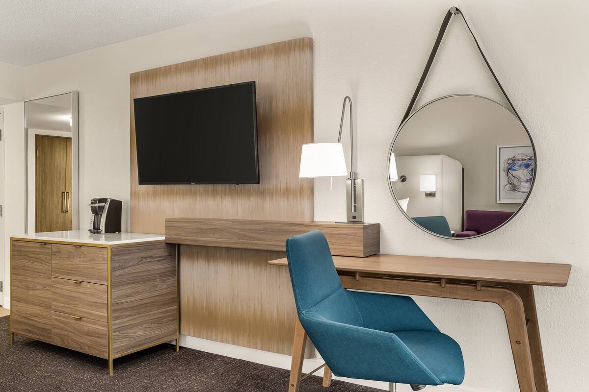 Ein Hotelzimmer mit einem Holztisch, einem blauen Sessel, einem Flachbildfernseher, einem Spiegel und einer Lampe.