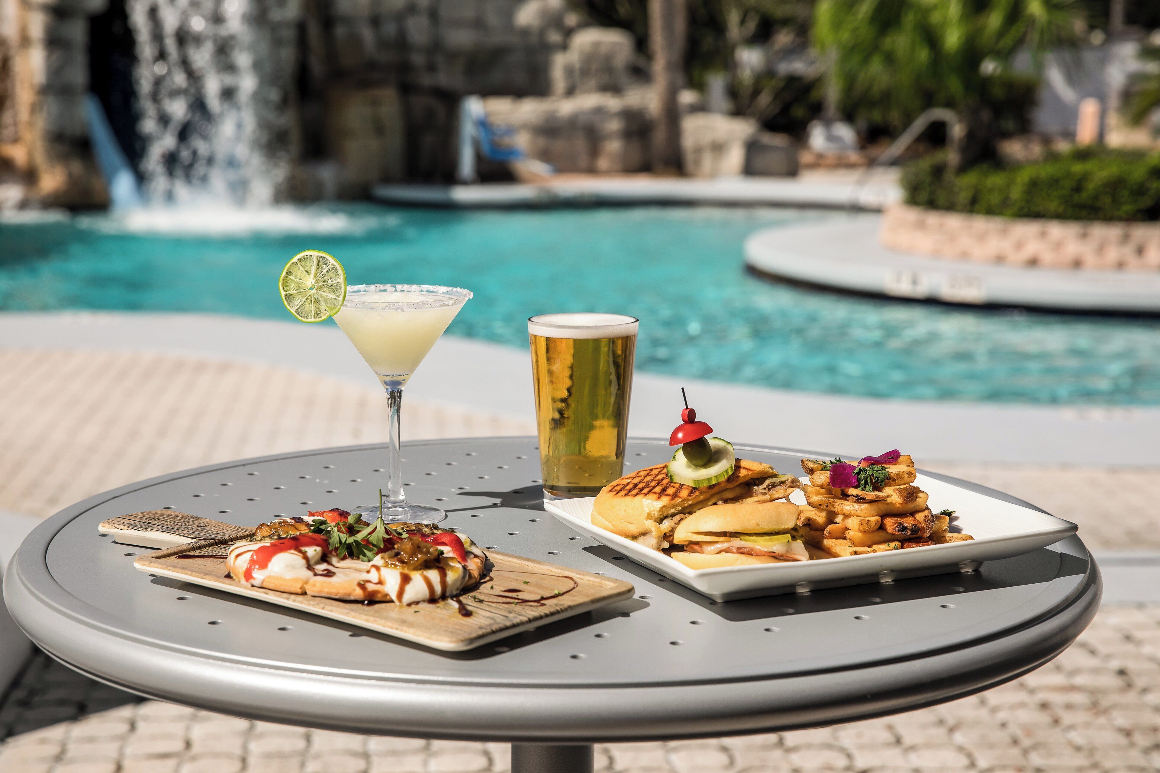A table with a meal consisting of grilled sandwiches and fries, a cocktail with a lime garnish, and a beer beside a pool with a waterfall in the background.