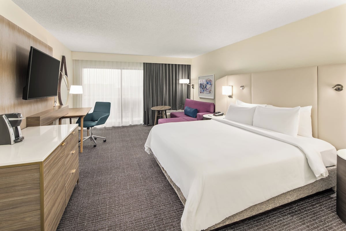 Ein Hotelzimmer mit einem großen Bett, einem Schreibtisch mit Flachbildfernseher, einem Sessel, einem kleinen Tisch und einem Fenster mit Vorhängen.