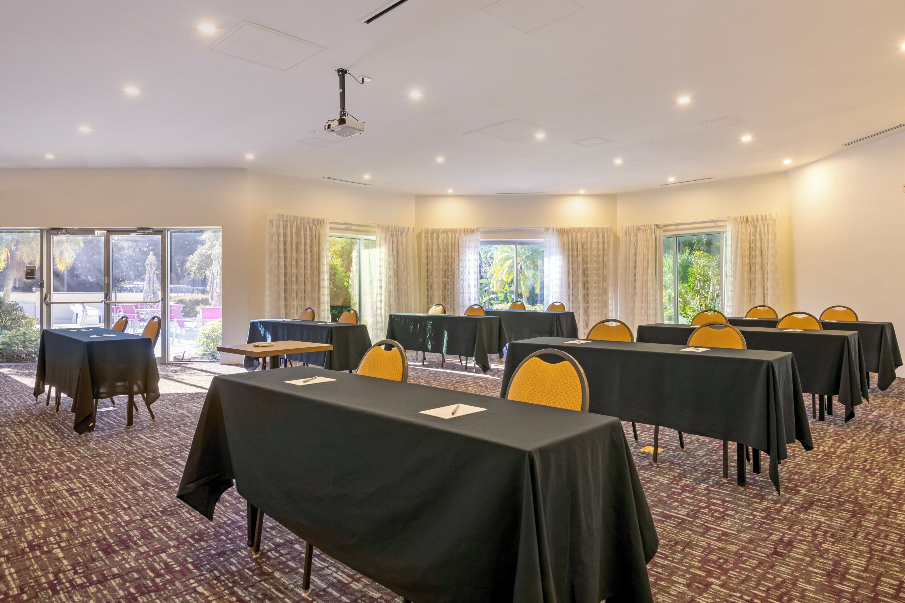 Un salón de conferencias con mesas y sillas, ventanas amplias y piso alfombrado.