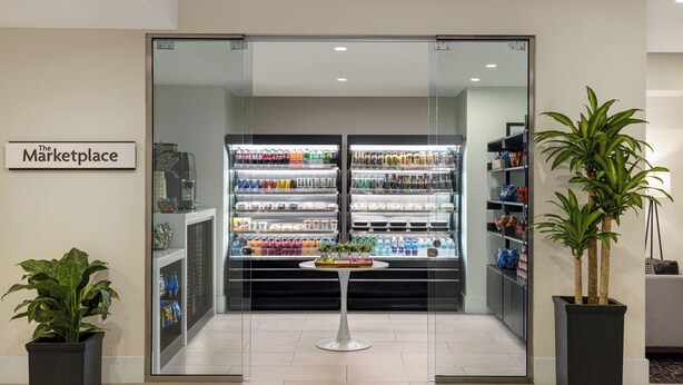 A modern interior with a glass door entrance labeled "The Marketplace," featuring refrigerated beverage dispensers and shelves stocked with snacks. - Crowne Plaza Orlando - Lake Buena Vista  by IHG (Orlando)