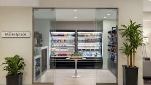 Un intérieur moderne avec une entrée en verre marquée de l’inscription « The Marketplace », comprenant des distributeurs de boissons réfrigérés et des étagères remplies de collations.