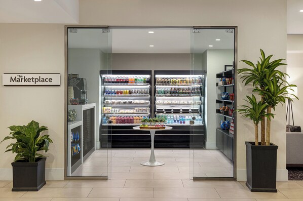 A modern interior with a glass door entrance labeled "The Marketplace," featuring refrigerated beverage dispensers and shelves stocked with snacks. - Crowne Plaza Orlando - Lake Buena Vista  by IHG (Orlando)