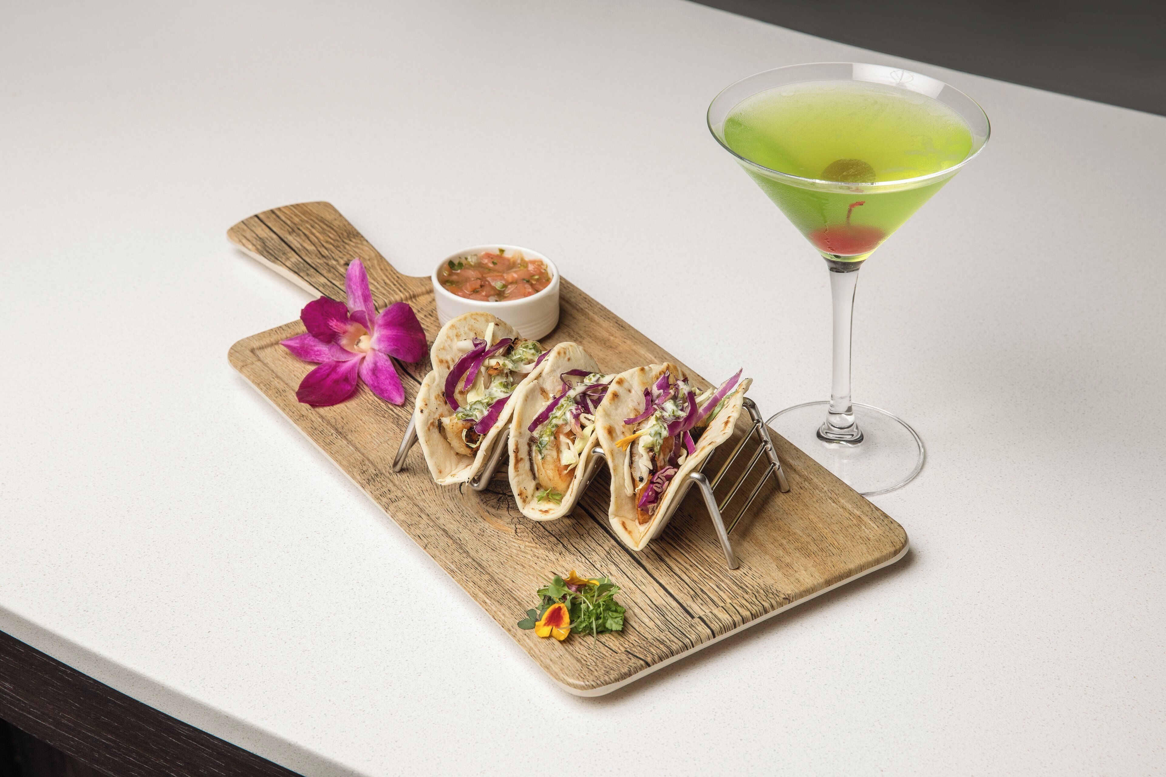A wooden board with three tacos, a small bowl of salsa, and a martini glass with a green drink.