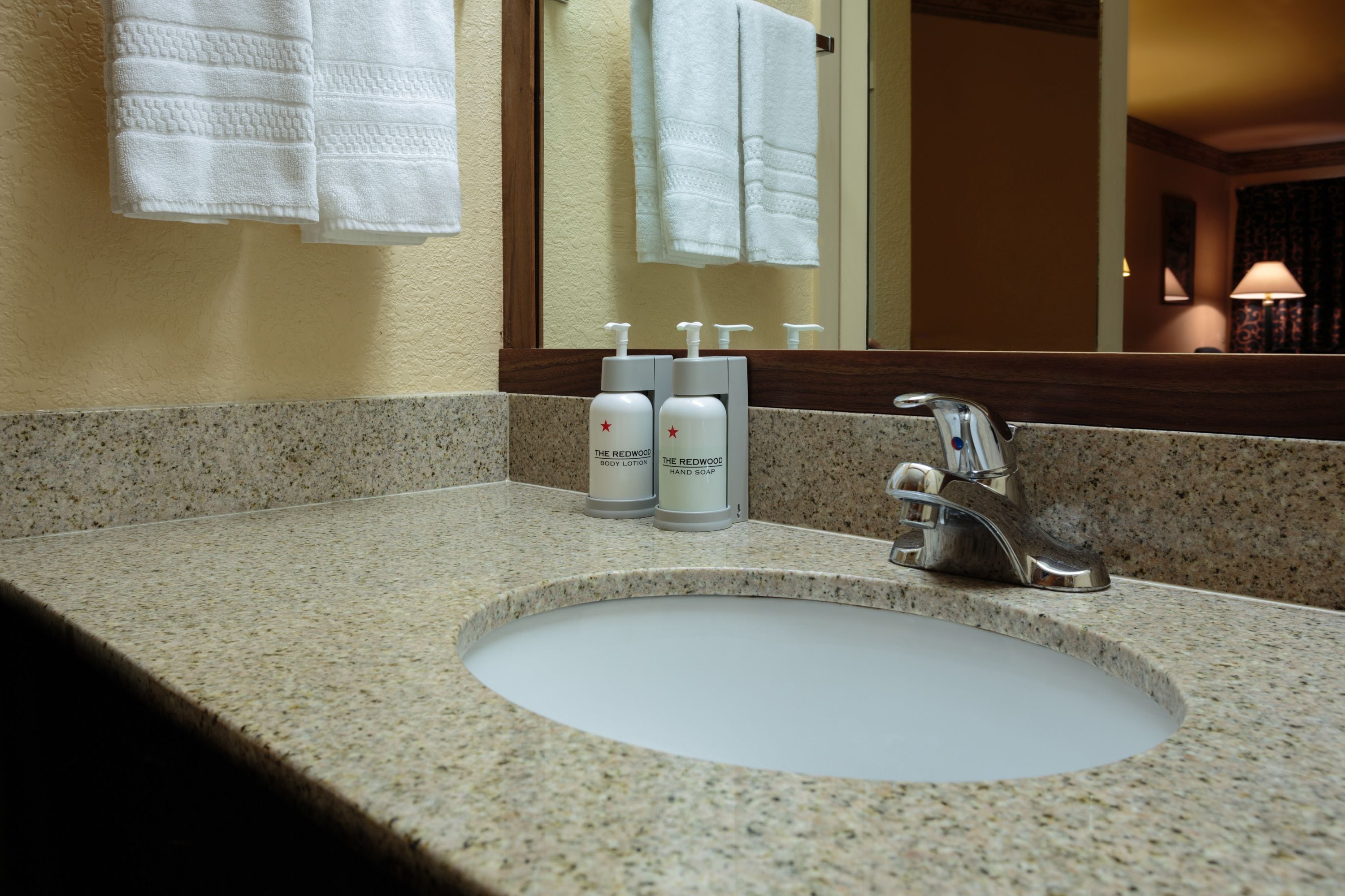 A bathroom sink with a granite countertop, two bottles of The Redwood body lotion, and a mirror above the sink.