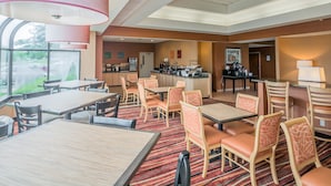 A hotel dining area with tables and chairs, a kitchen in the background, and large windows. - Comfort Inn - NYS Fairgrounds (Syracuse)