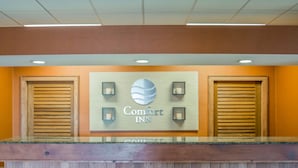 A reception desk at Comfort Inn with a sign, two wooden shutters, and three wall-mounted lights. - Comfort Inn - NYS Fairgrounds (Syracuse)