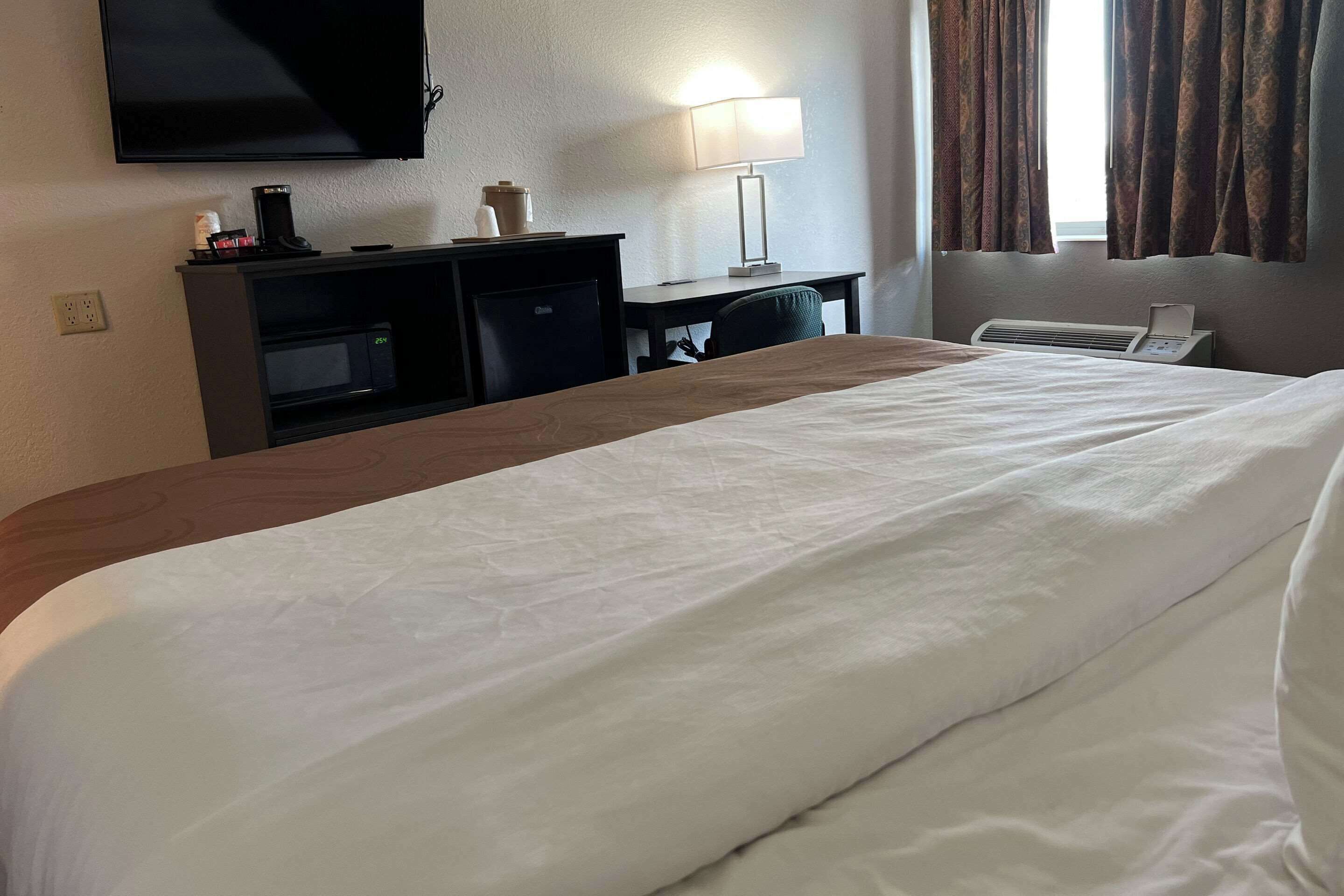 A hotel room with a large bed, a desk with a lamp, a television, and a window with curtains.