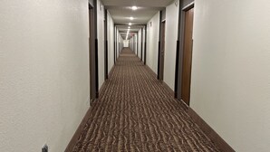 Un long couloir étroit, typique des hôtels, avec un tapis à motifs, des murs blancs et des portes en bois.